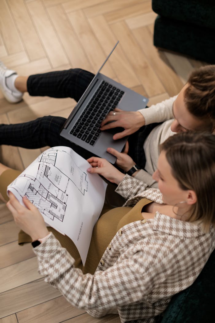 A couple sits on the floor examining house blueprints and using a laptop, planning their future home.